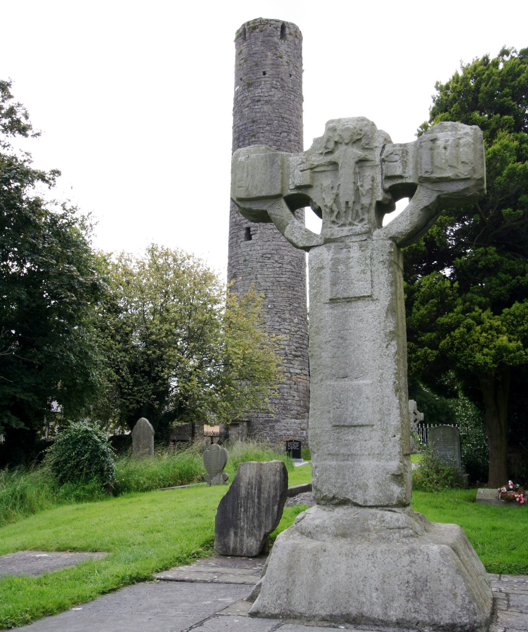 [Image: Most of a stone Celtic cross, some grave stones in a green lawn, and in the background some trees and a tall, thin stone tower.]